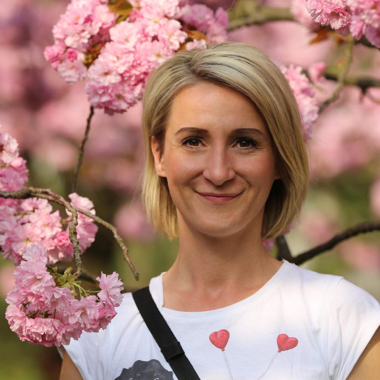 Franziska, Tiersitterin für Katzen und kleine Haustiere in Babelsberg Eine Tiersitterin mit blonden Haaren steht vor blühenden Kirschbäumen. (Portrait)